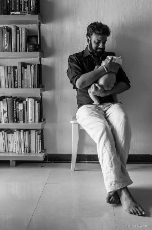 Father with his infant son sitting on a stool next to a shelf full of books.